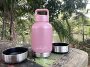 A dusty pink insulated water bottle with a matching pink lid handle sits on a stone surface outdoors, with two stainless steel lids placed beside it. Trees and a body of water are blurred in the background.