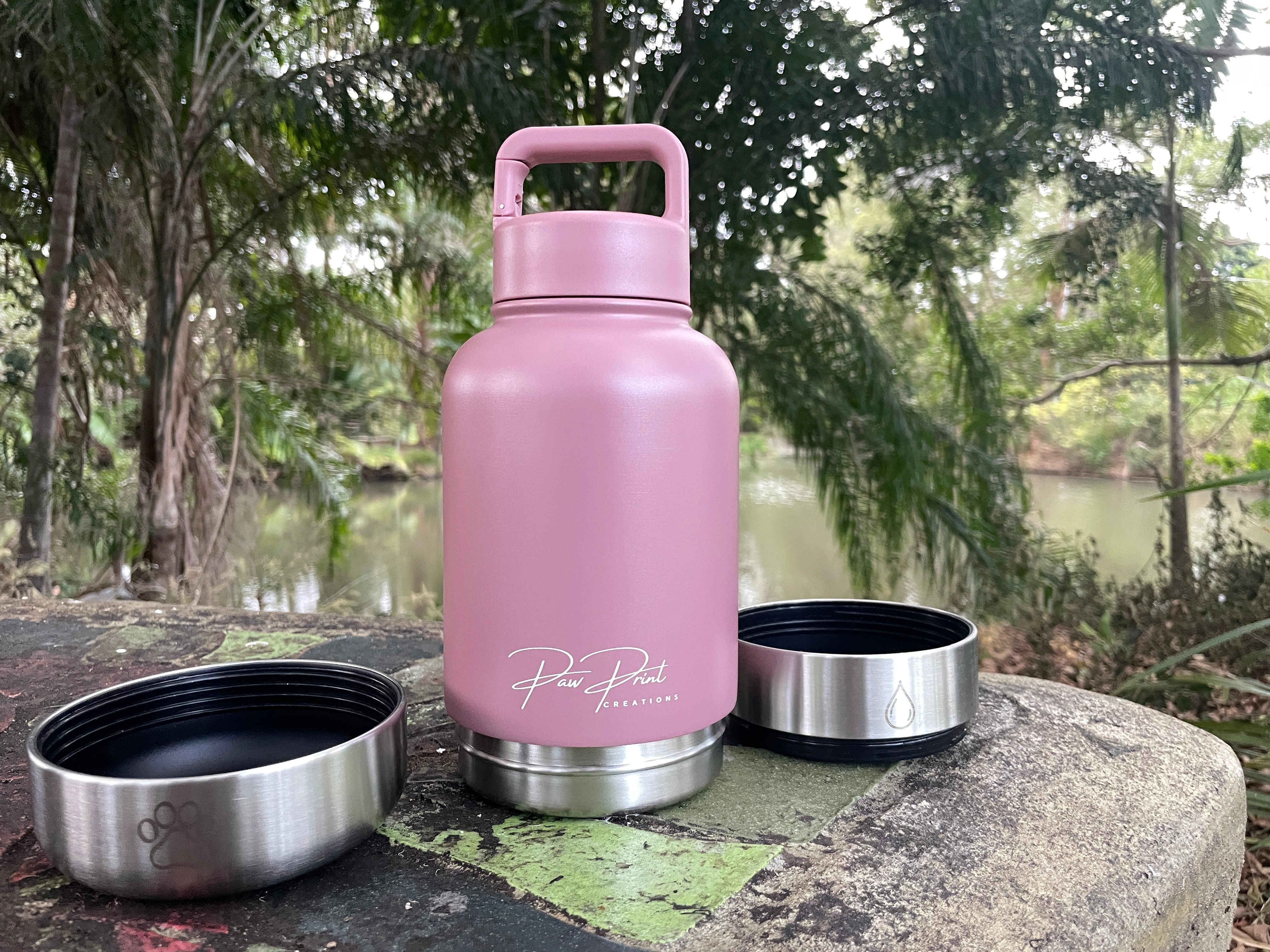 A dusty pink insulated water bottle with a matching pink lid handle sits on a stone surface outdoors, with two stainless steel lids placed beside it. Trees and a body of water are blurred in the background.