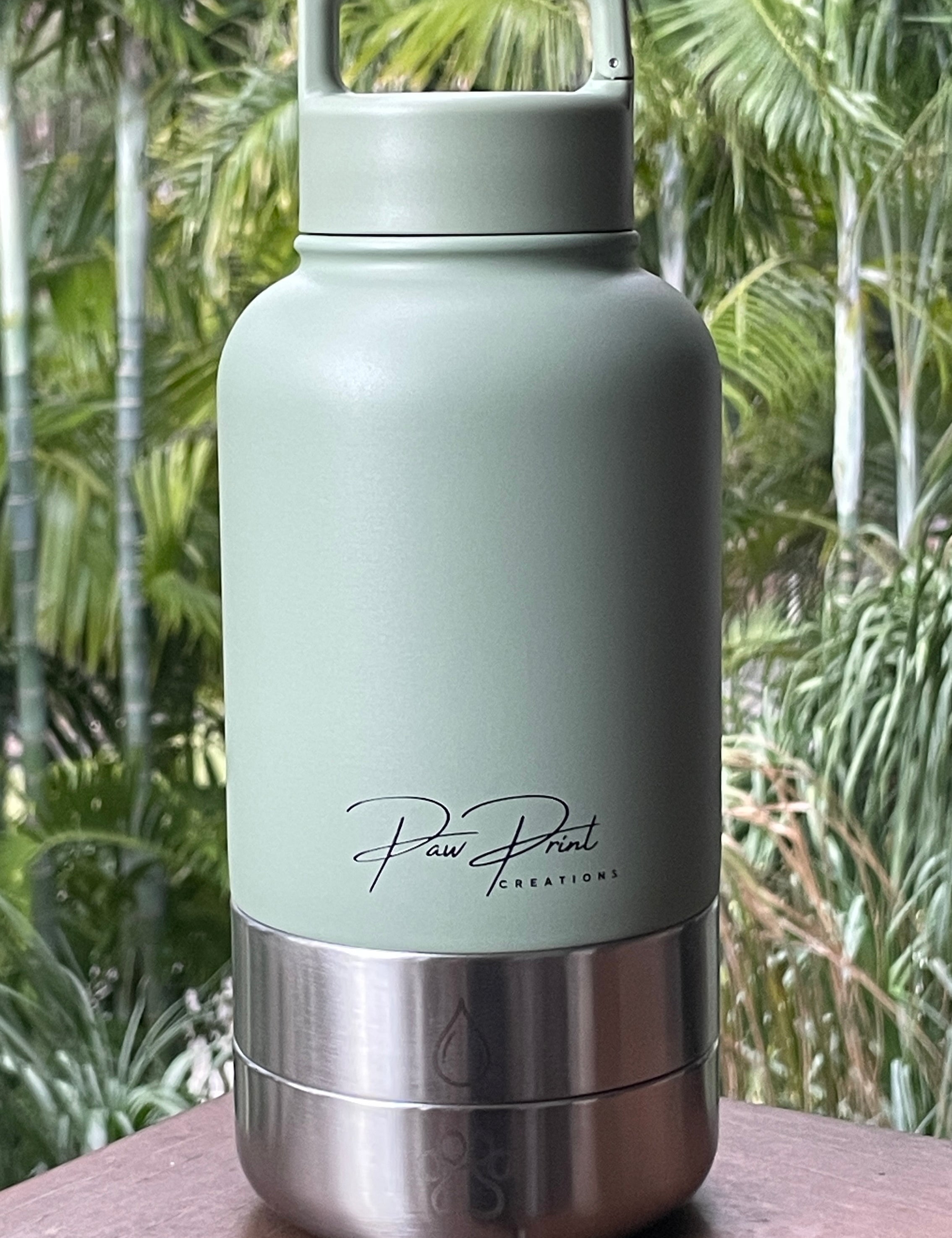 A matte sage green insulated water bottle with a stainless steel base, resting on a wooden surface with a blurred tropical garden background. The brand name 'Paw Print Essentials' is printed in black script on the front.