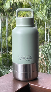 A matte sage green insulated water bottle with a stainless steel base, resting on a wooden surface with a blurred tropical garden background. The brand name 'Paw Print Essentials' is printed in black script on the front.