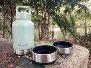 A pale mint green insulated water bottle with a carry loop stands on a stone outdoors, next to two stainless steel lids with black interiors. Trees and a body of water are blurred in the background.