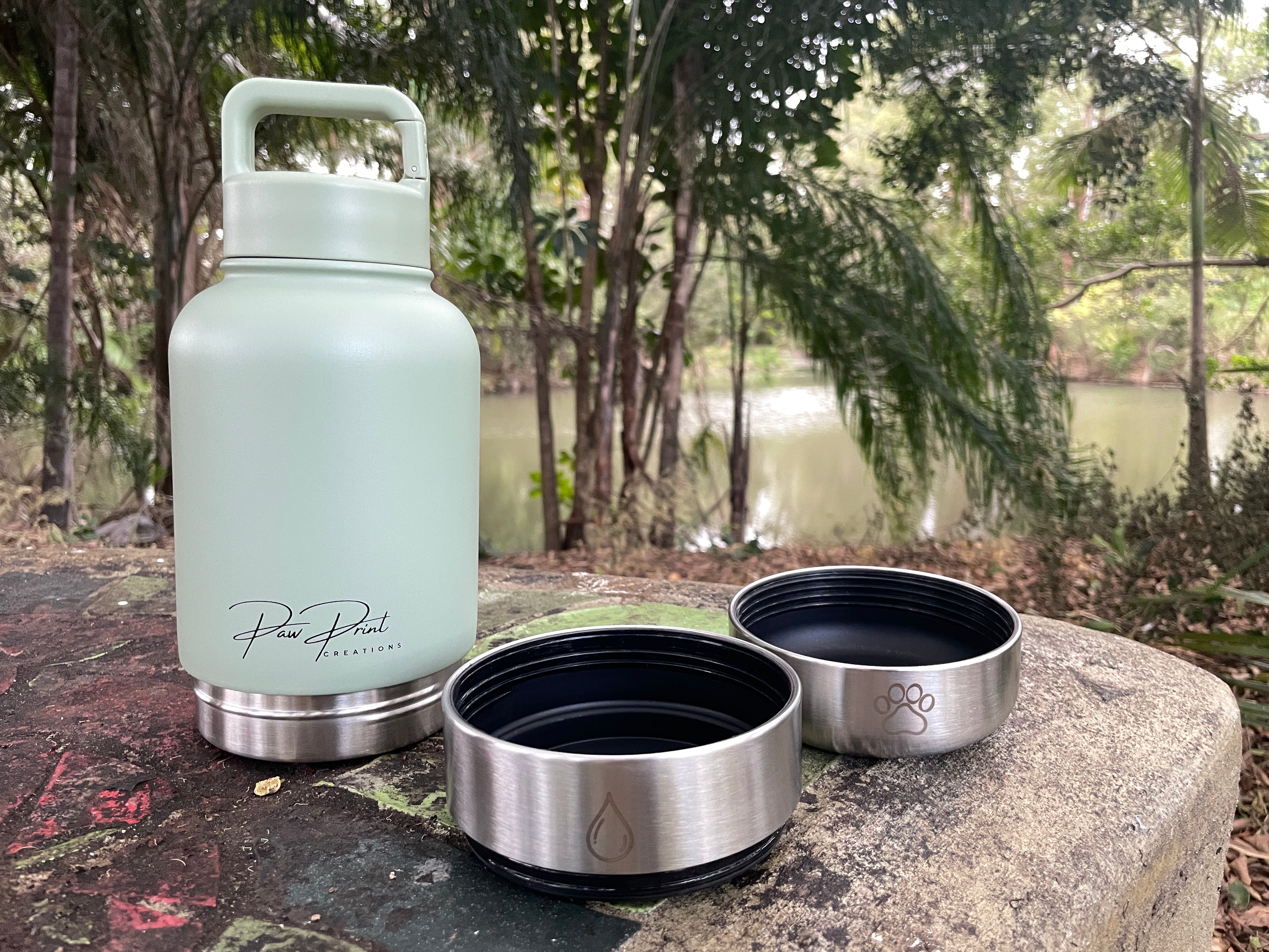 A pale mint green insulated water bottle with a carry loop stands on a stone outdoors, next to two stainless steel lids with black interiors. Trees and a body of water are blurred in the background.